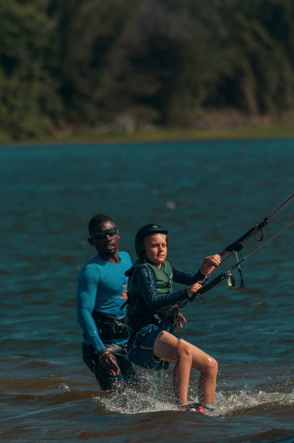 A young student up on the board and riding, Wendell standing in the lagoon right behind